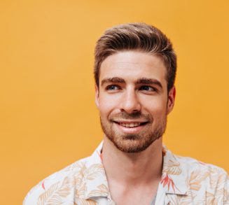 Portrait of nice young man with stylish hairstyle, blue eyes and beard in light cool shirt looking Portrait of nice young man with stylish hairstyle, blue eyes and beard in light cool shirt looking away and smiling on orange backdrop..