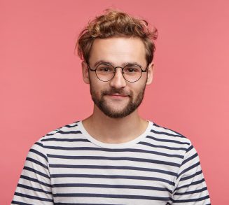 Indoor shot of pleasant looking bearded hipster guy wears spectacles and striped t shirt, looks directly into camera, isolated over pink studio background. Male student spends weekends at home alone Indoor shot of pleasant looking bearded hipster guy wears spectacles and striped t shirt, looks directly into camera, isolated over pink studio background. Male student spends weekends at home alone